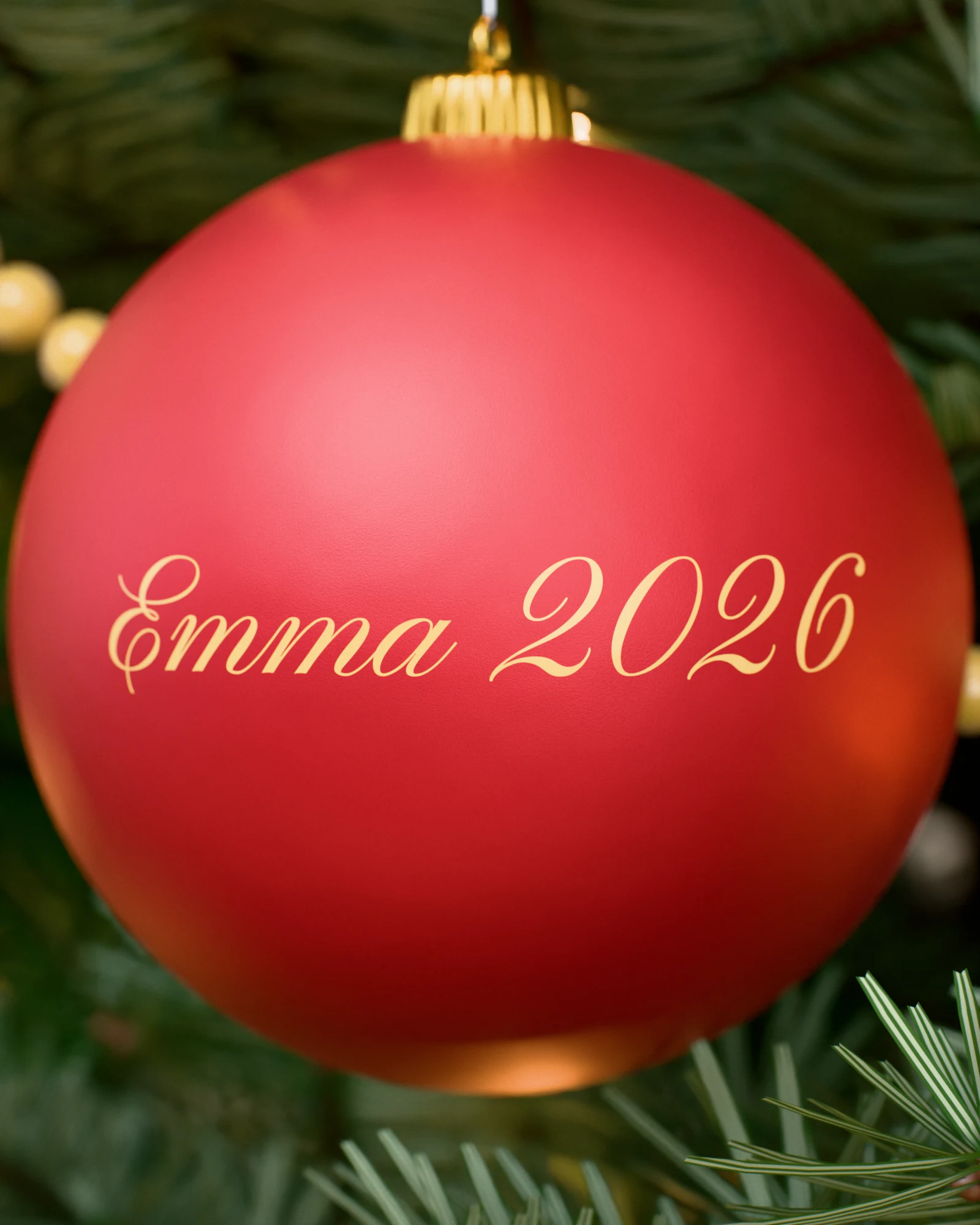 Matt red bauble with “Emma 2026” in gold script lettering among fir branches